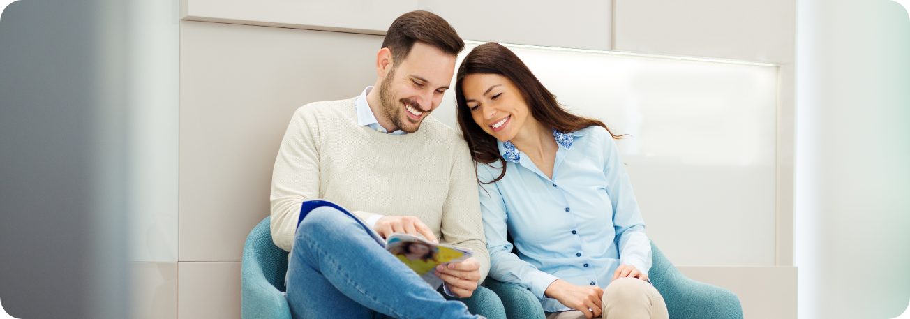 A smiling couple sitting in a modern dental clinic waiting area, reading a brochure about dental services.